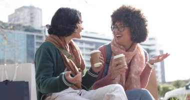 Bayan arkadaşlar kahve içerken selamlaşıp sohbet ediyorlar, alışveriş torbalarıyla parkta oturuyorlar. Gündelik, dostluk, rahatlama, açık hava, şehir, yaşam tarzı, neşeli