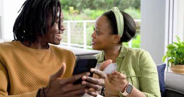 Afro-Amerikalı çift akıllı telefonları dinliyor, kanepede kart tutuyorlar, onaylandıktan sonra beşlik çakıyorlar. İlişki, samimiyet, rahatlama, konforlu, teknoloji, ev, bağlantı