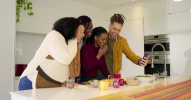 Çeşitli arkadaşlar akıllı telefonları seçiyor ve mutfak adasında selfie çekiyorlar sonra da salata hazırlıklarına devam ediyorlar. Sosyalleşme, yaşam tarzı, modern, canlı, gündelik, arkadaşlık, yemek pişirme