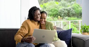 Kredi kartına tepki veren Afro-Amerikalı çift gri kanepede, dizüstü bilgisayardaki alışverişi kutluyorlar. Romantik, samimi, modern, yaşam tarzı, teknoloji, mutluluk, bağlantı