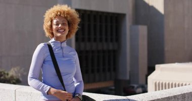 Afrika kökenli Amerikalı bir kadın çatıdaki korkuluklara yaslanmış güneşi hissediyor ve spor çantasını tutarken gülüyor. Kentsel, yaşam tarzı, çağdaş, fitness, motivasyon, boş zaman, enerjik