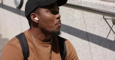Afro-Amerikalı bir adam kulaklıklarla dışarıda yürüyen merdivenleri çiğniyor, gün ışığına doğru dönüyor ve gülümsüyor. Kentsel, gündelik, rahat, güneş ışığı, düşünceli, açık hava, modern