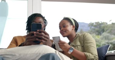 Kanepeden satın almak için kart detaylarını girmeden önce akıllı telefondan Bill 'e bakan Afro-Amerikalı çift. Teknoloji, iletişim, rahatlama, ev, yaşam tarzı, dijital, bağlantı