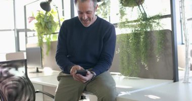 Orta yaşlı bir adam hoş bir uyarı aldıktan sonra modern ofis masasında akıllı telefon kaydırıyor. Profesyonel, çalışma alanı, modern, teknoloji, iletişim, rahatlama, üretkenlik