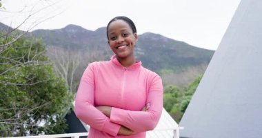 Pembe ceketli Afro-Amerikalı kadın gülerek ve terasta korkuluklara yaslanarak izleyicileri davet ediyor. Açık hava, manzara, kendine güven, rahatlık, yaşam tarzı, çağdaş doğa.