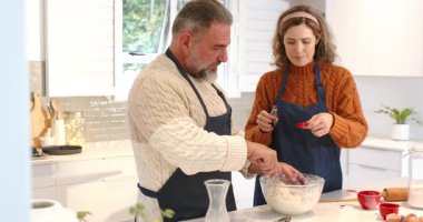 Mutfak adasında cam kasede hamuru karıştıran bir çift, fırında pişirmek için baharat serpiyor ve ölçüyor. Aile içi, aşçılık, yaşam tarzı, kırsal, sıcak, takım çalışması, sade