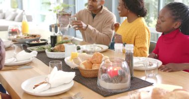 Olgun bir adam su sürahisini kaldırırken çeşitli aileler içkileri doldurur ve sofrada yemek hazırlar. Aile toplantısı, kutlama, neşe, sıcak atmosfer, kırsal, birliktelik, açık hava ışığı.