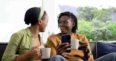 Afro-Amerikalı çift kanepede şaklabanlık ve kaydırma akıllı telefonlarıyla gülüyor, fincanları yudumluyorlar. Romantizm, konfor, bağlantı, rahatlık, yakınlık, yaşam tarzı, kırsal