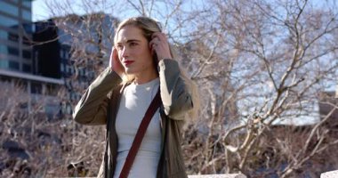 Beton parmaklıklarda duruyor, 20 'li yaşlarda bir kadın müzik dinlemek için kulaklık kaldırıyor ve çanta tutuyor. Şehir, huzur, yaşam tarzı, modern, rahatlama, teknoloji, açık hava