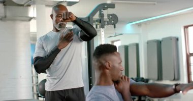 Jimnastik başlangıcında, Afrikalı Amerikalı antrenör müşteriye hizalama için göğüs pres makinesine rehberlik ediyor. Dayanıklılık eğitimi, fitness, egzersiz, hareketlilik, esneklik, kas aktivasyonu, fizik tedavi