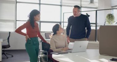 Tekerlekli sandalyedeki kadın ofiste dizüstü bilgisayarda yazmaya başlıyor, iş arkadaşları katılıyor, ekranı işaret ediyor. İşbirliği, takım çalışması, çeşitlilik, erişilebilirlik, modern, ofis, iletişim