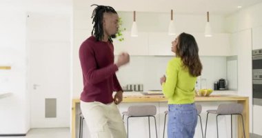 Afro-Amerikalı erkek ve Hintli kadın dayak duyuyor, mutfak adasının yanında dans ediyor, köpek okluyor. Kutlama, sevinç, bağlantı, hareket, ev, yaşam tarzı, çağdaş