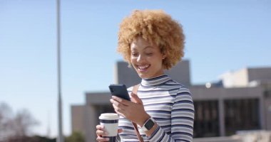 Bir kadın telefona bakıyor, akıllı telefonun kilidini açıyor ve plazada elinde kahveyle gülümseyen mesajları kontrol ediyor. Gündelik, açık hava, yaşam tarzı, modern, bağlantı, boş zaman, şehir