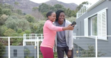 Siyah çift akıllı telefon kullanıyor, selfie çekiyor, çekim yapıyor ve balkonda gülüyor. Romantizm, açık hava, eğlence, yaşam tarzı, samimiyet, yeşillik, şehir