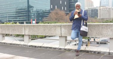 Kıdemli Afro-Amerikan adam bariyerin önünde durmuş sandviç yiyor akıllı telefon, kopyalama uzayı. Kentsel, yaşam tarzı, samimi, modern, düşünceli, sıradan, boş zaman