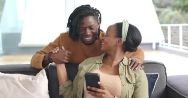 Afro-Amerikalı çift mesajlaşmaya, yumruk tokuşturmaya ve kanepede el ele tutuşup akıllı telefon seyretmeye tepki veriyor. Romantizm, şefkat, bağlantı, samimiyet, konforlu, modern, yaşam tarzı