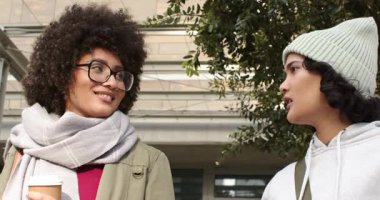 Asyalı bir arkadaş, kahve fincanı tutan Afro-Amerikalı bir kadınla konuşuyor. Yapraklı tepe örtüsünün altında yürüyor. Arkadaşlık, arkadaşlık, gezinti, günlük konuşma, şehir, yeşillik.