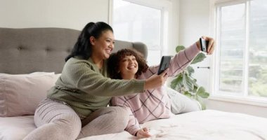 Anne-kız selfie çekiyor, akıllı telefonları inceliyor ve yatakta oturuyorlar. Aileler, bağlılık, arkadaşlık, memnuniyet, teknoloji, yaşam tarzı, konforlu