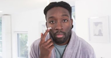 Afro-Amerikalı bir adam kameraya bakıyor, cilt bakımına başlıyor, lavabonun altında nemlendirici sürüyor. Cilt bakımı, kendine bakım, sağlık, güzellik, tımar, doğal ışık, minimalizm.
