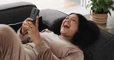 Koyu gri kanepede uzanan orta yaşlı bir kadın konsolu kaldırıyor ve pencere kenarında oyun oynarken gülüyor. Rahatlama, eğlence, rahatlık, konfor, teknoloji, gündelik, mutluluk