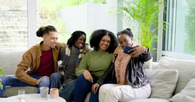 Dört arkadaş akıllı telefon yetiştiriyor, gülüyor ve evdeki kanepede selfie çekerken ekrana dokunuyor. Arkadaşlık, birliktelik, rahatlama, modern, samimi, neşeli, yaşam tarzı