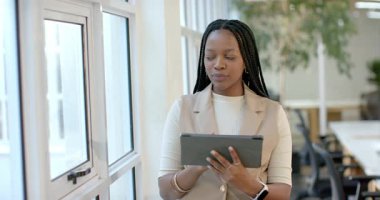 Afro-Amerikalı bir kadın içeriği gözden geçirmek için tablet ekranına tıklıyor, ofiste gülümseyen pencerenin önünde duruyor. Profesyonel, teknoloji, modern, çalışma alanı, yenilik, üretkenlik, kadın