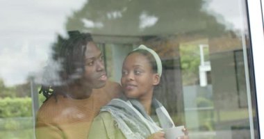 Afro-Amerikalı bir çift cam kenarında durup, kupa tutuyor, bahçeye bakıyor, konuşuyor ve işaret ediyor. Romantik, rahat, düşünceli, modern, doğal, samimi, yaşam tarzı