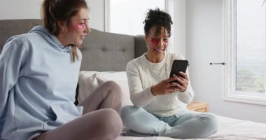 Çeşitli bayan arkadaşlar yatağa uzanıp akıllı telefonları dinliyorlar, göz bandı takarak selfie çekiyorlar. Arkadaşlık, rahatlama, kişisel bakım, konforlu, teknoloji, yaşam tarzı, rahatlık