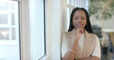 Pencerenin yanında, bej ceketli Afro-Amerikalı kadın poz verirken ofisteki kahkahalara geçiyor. Profesyonel, kentsel, samimi, açık görüşlü, çağdaş, neşeli, kendinden emin.