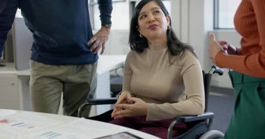 Tekerlekli sandalyedeki tableti işaret eden iş arkadaşı olarak çeşitli iş arkadaşları ofiste verileri tartışıyor. İşbirliği, takım çalışması, analitik, modern, kapsayıcı, üretkenlik, iletişim