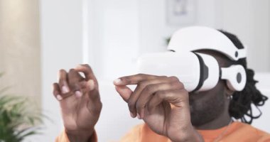 Afro-Amerikan erkek, VR kulaklık takıyor ve arayüzü kullanmak için oturma odasındaki kontrolleri manipüle ediyor. Gelecekçi, teknoloji, etkileşimli, sürükleyici, jest, sanal gerçeklik, modern