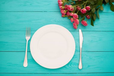 Top view of tableware of round and heart shaped empty plate, metal knife and fork served on white wooden table with glowing candle and gift box prepared for celebration of saint valentine day