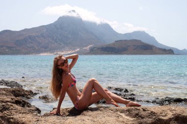 Kadın mayo giymiş. Sahilde ve dağlarda poz veriyor.