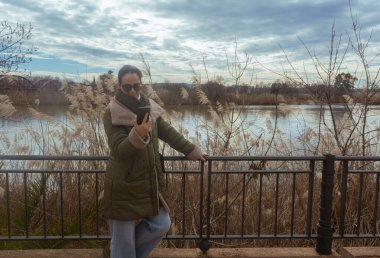 Adult woman looking at cell phone screen, on a walk along river clipart