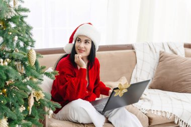 Noel Baba şapkalı güzel bir kız Noel ağacının yanında oturuyor ve Noel için ne alacağını düşünüyor. İnternetten online alışveriş. Yılbaşında dizüstü bilgisayarı olan bir kız..