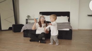 Anne ve oğlu sabun köpüğüyle oynuyorlar. Çocuk sabun köpüklerinin tadını çıkarıyor. Aile evde oturup sabun köpüğüyle oynuyor. Mutlu bir çocukluk ve sağlıklı bir aile, mutlu aile ilişkileri.