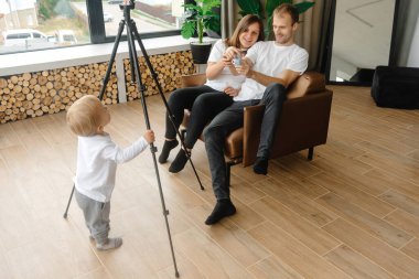 Bir çocuk blogcu elinde bir video tripod tutuyor ve genç bir blogcu olmaya hazırlanıyor. Ebeveynler bir sandalyede yan yana oturuyorlar.