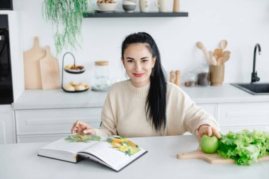 İyimser genç bir kadın kameraya bakar ve mutfakta otururken gülümser. Sağlıklı beslenme ve yaşam tarzıyla ilgili bir dergi okuyorum..