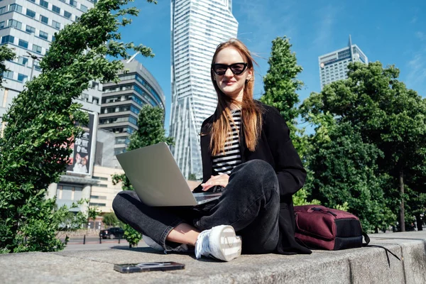 Young woman student, freelancer with backpack and laptop sitting on modern downtown high-rises skyscrapers street city view background. Female worker with modern backpack and laptop in megapolis
