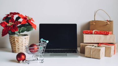 E-Ticaret 'te başarılı bir Noel sezonu, online Noel alışverişi. Dizüstü bilgisayarı aç Ekran, mini alışveriş arabası, Poinsettias Noel Çiçeği ve beyaz masadaki hediye kutuları.