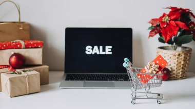 E-Ticaret 'te başarılı bir Noel sezonu, online Noel alışverişi. Dizüstü bilgisayarı aç Ekran, mini alışveriş arabası, Poinsettias Noel Çiçeği ve beyaz masadaki hediye kutuları.