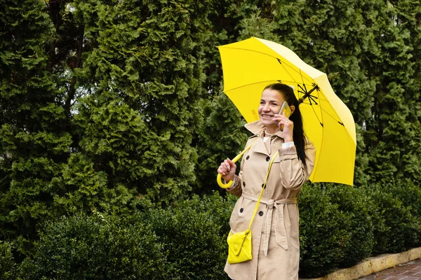 Happy senior woman in yellow rain coat with yellow umbrella is talking on a smartphone and walking in park. Staying in touch with distant loved ones.
