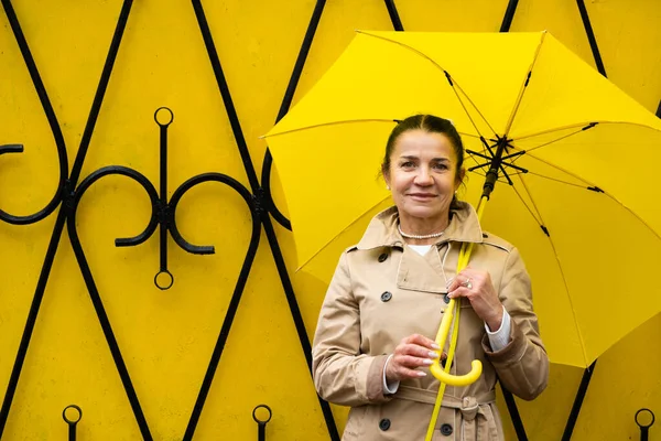 Stress resilience and mental health, concept. Managing stress and building resilience. Happy senior woman in yellow rain coat with yellow umbrella walking in park