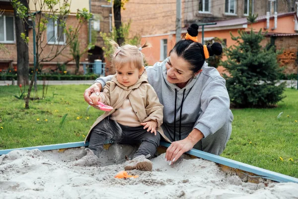 Benefits of Sand Play. Sandpit play ideas to get kids learning while having fun. Senior woman grandmother and little toddler girl granddaughter together playing in sandbox in spring garden.