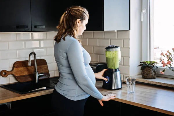 为怀孕和产后 产前营养提供的绿色平滑疗法 怀孕妇女在厨房里用搅拌机和饮料配制绿色维生素冰沙 — 图库照片