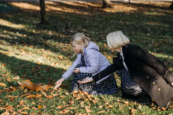 Happy funny mom and teen daughter feeding nuts to a squirrel on autumn park on a sunny day. Family spend time together and enjoy autumn fall season.
