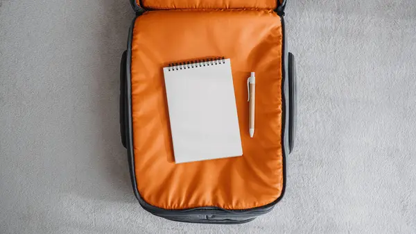 旅行パッキング バケーションのモックアップのためのリスト 旅行のパッキング チェックリスト 休日またはビジネス旅行のため 自宅でベッドのオレンジのスーツケースにペンが付いている空のノートパッドを開けて下さい — ストック写真