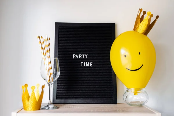 Party time fun celebration event minimal concept with smiling yellow balloon with golden crown, blackboard with text Party time and party stuff on white.