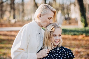 Anne ile kızı arasında duygusal bir an yaşandı. Sonbahar yapraklarıyla dolu güneşli bir parkta kızının başını öpen bir anne sahnesi.