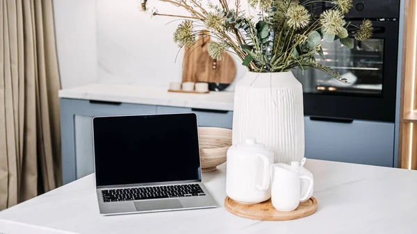 Home Office Setup with Laptop and Cozy Kitchen Decor. A chic home office setup featuring an open laptop on a marble countertop, complemented by a warm kitchen ambiance and stylish decor